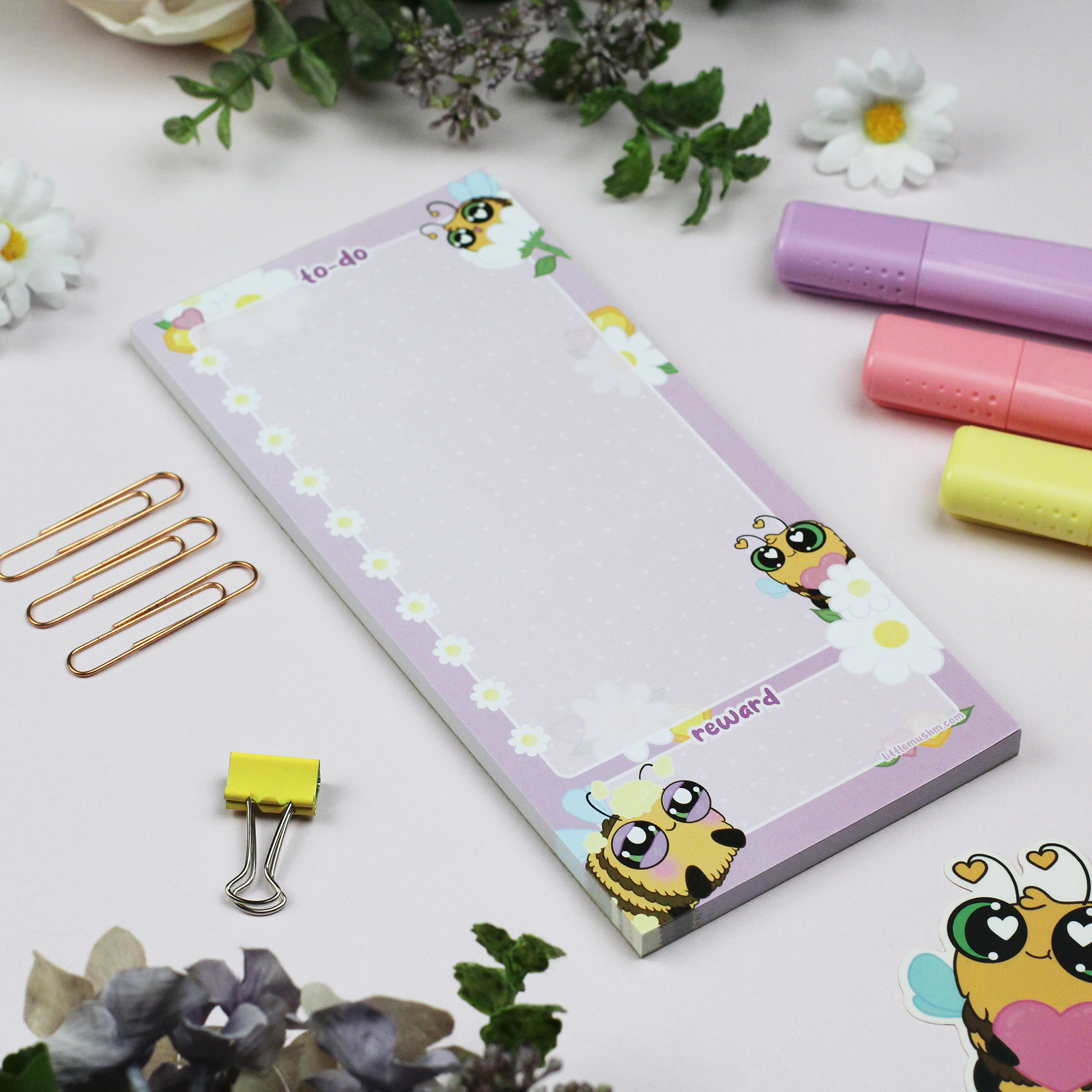 A notepad with a cartoon bee design, titled 'To-Do' with daisy checkboxes and a reward section, accompanied by a purple pen, yellow highlighter, and copper-colored pencils on a floral background.